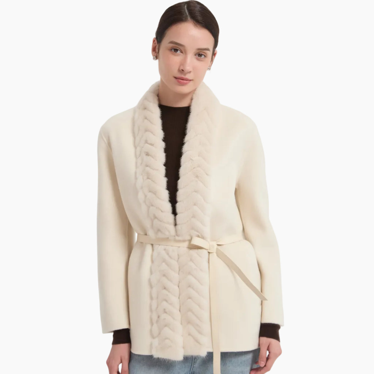 A woman modeling a cream-colored wrap coat with a thick faux-fur collar and a matching tie belt, worn over a dark brown top and paired with light blue jeans, against a plain light gray background.