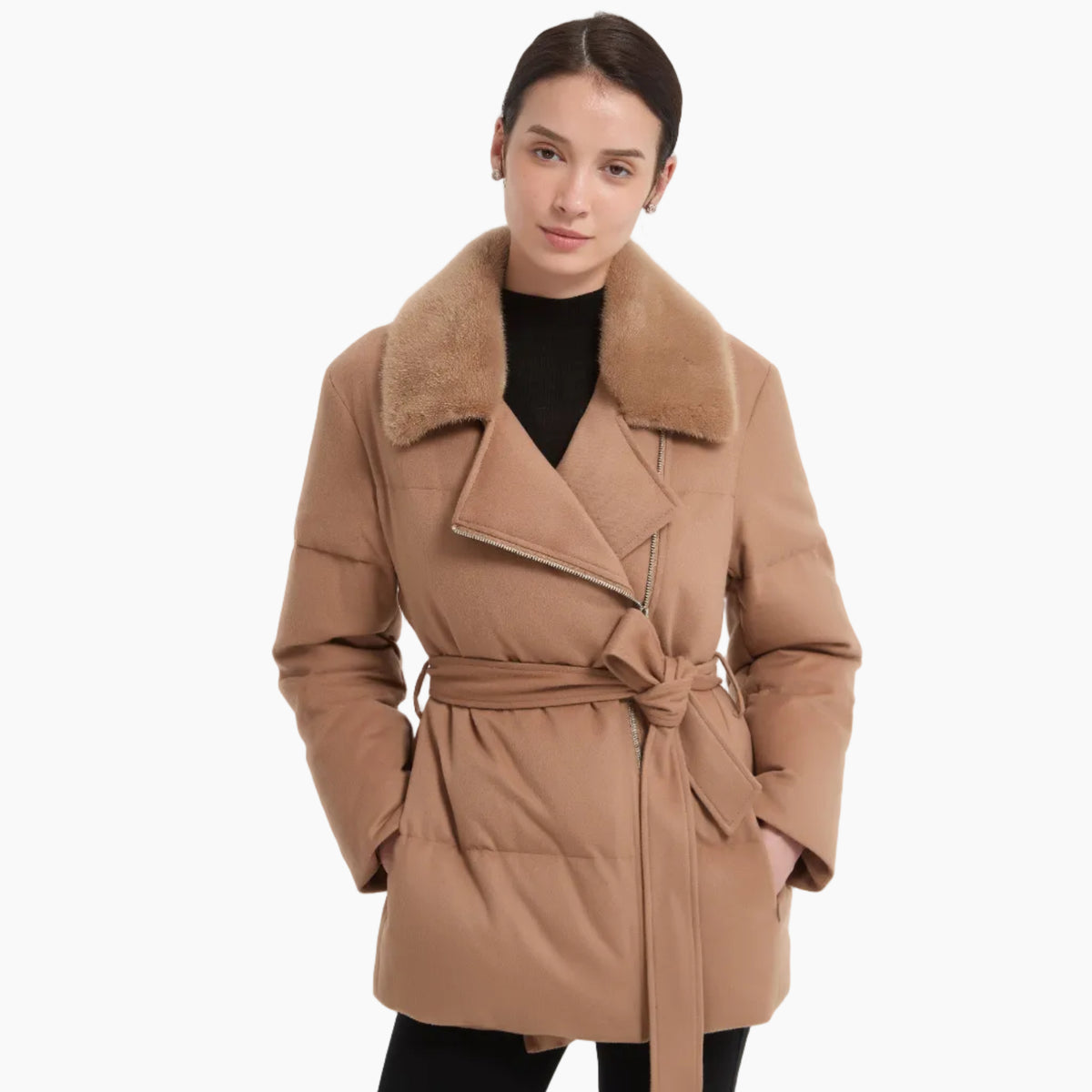 A woman modeling a camel-colored puffer coat with a plush faux-fur collar, belted at the waist, worn over a black top and black pants against a light gray studio background.