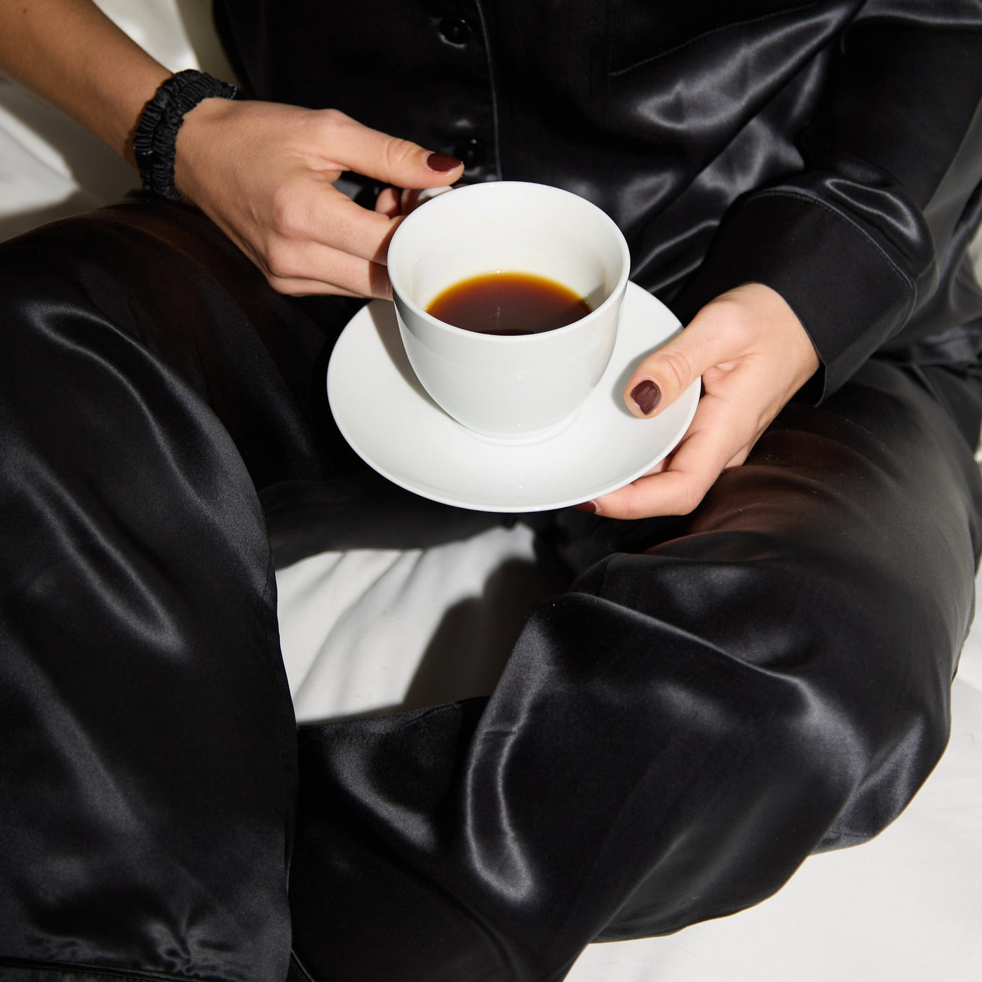 Person in black silk pajamas holding a white cup of coffee.