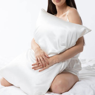 Woman sitting on a bed holding a large ivory Silk Works mulberry silk pillowcase, dressed in ivory cami sleep set