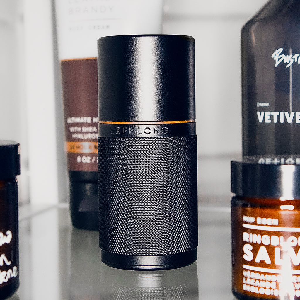 Black refillable deodorant with knurled texture, bronze detail and logo saying Lifelong. Around, personal care products in bottles.