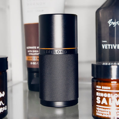 Black refillable deodorant with knurled texture, bronze detail and logo saying Lifelong. Around, personal care products in bottles.