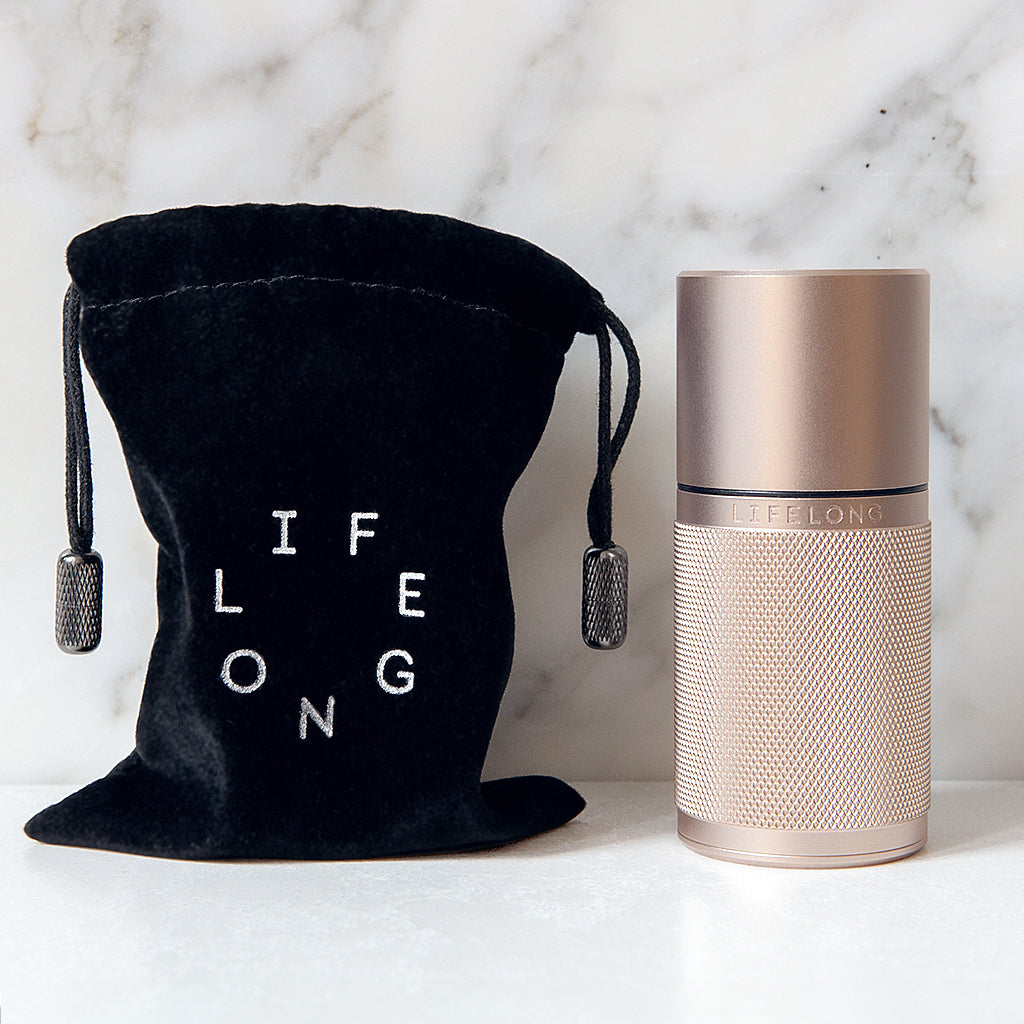 Rose-coloured refillable deodorant with knurled texture, dark details and Lifelong logo. Beside, dark traveling bag with bright circled Lifelong logo. Background, white marble.