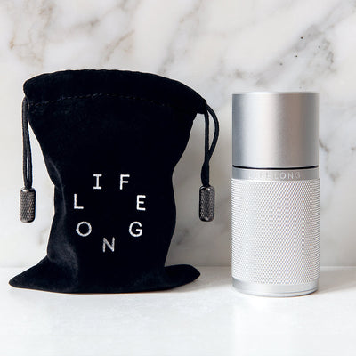 Silver-coloured refillable deodorant with knurled texture, dark details and Lifelong logo. Beside, dark traveling bag with bright circled Lifelong logo. Background, white marble.