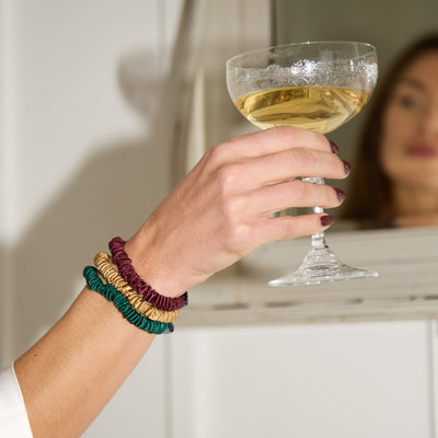Hand holding a glass of champagne with red plum, green and gold silk small scrunchies on a neutral background