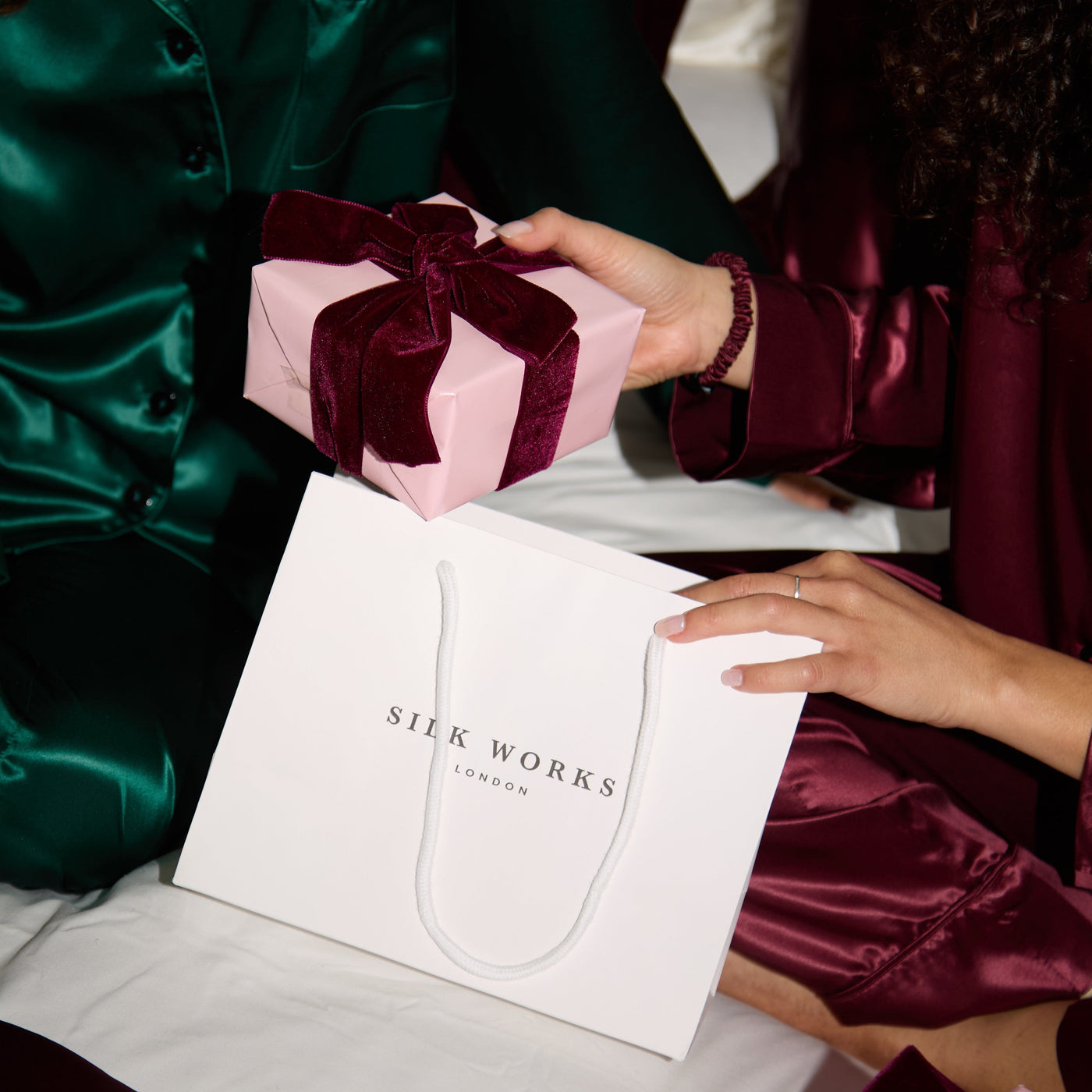 Person in green silk pyjamas outfit receiving a pink gift box with a bow from another person, with Silk Works London packaging.