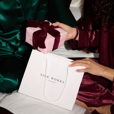 Person in green silk pyjamas outfit receiving a pink gift box with a bow from another person, with Silk Works London packaging.