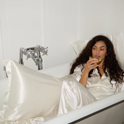 Woman in a white silk pyjamas sitting in a bathtub, holding a glass of champagne surrounded by silk pillowcases