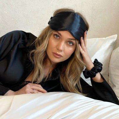 Woman reclining in bed wearing a Silk Works black mulberry silk eye mask and matching scrunchie, styled with silk sleepwear for a luxurious nighttime ritual.