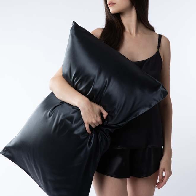 Woman in black silk sleepwear holding a Silk Works black mulberry silk pillowcase, styled against a clean white background.