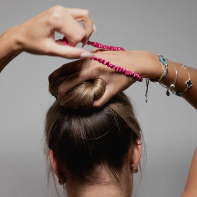 Woman styling her hair into a bun using a hot pink Silk Works mulberry silk scrunchie, showcasing gentle hold and frizz-free styling.