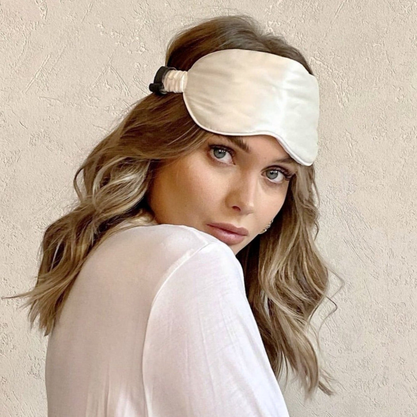 Woman wearing a Silk Works ivory mulberry silk eye mask with ruched strap, styled with soft waves and a white top, against a neutral textured background.