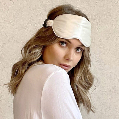 Woman wearing a Silk Works ivory mulberry silk eye mask with ruched strap, styled with soft waves and a white top, against a neutral textured background.