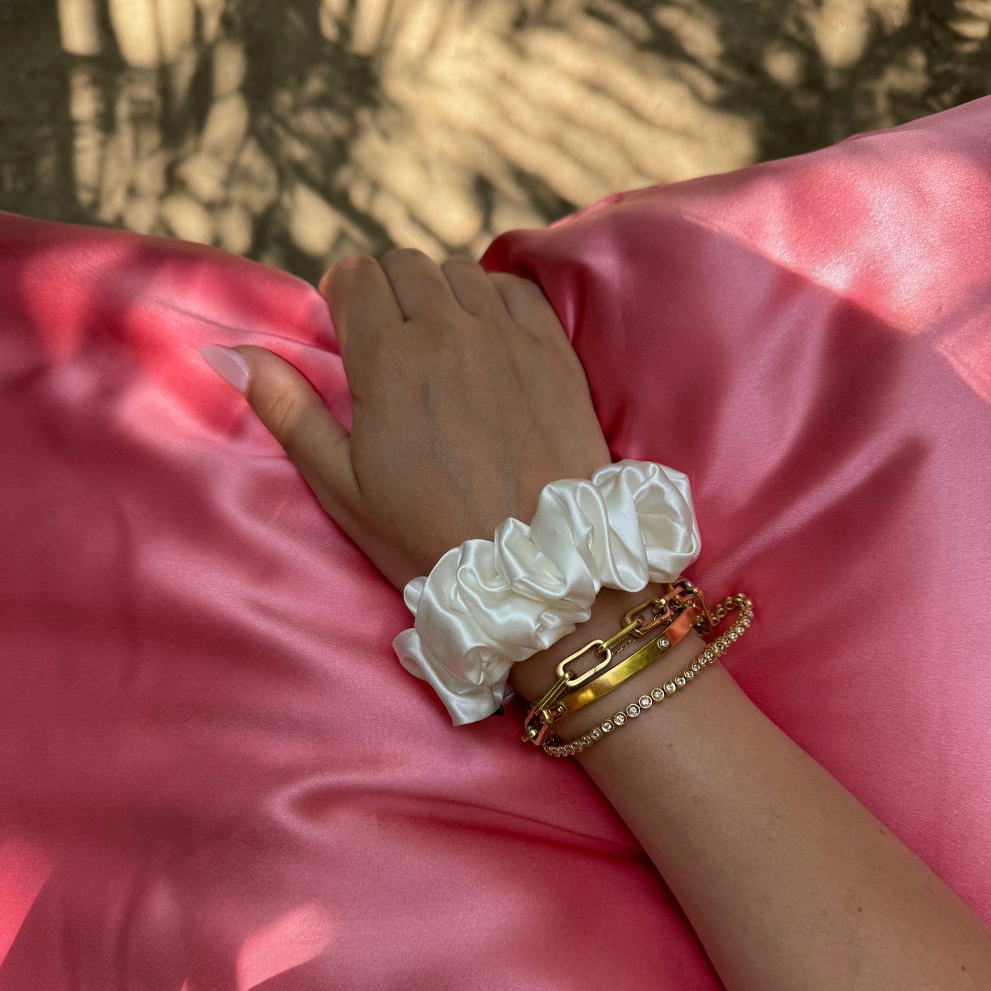 Hand adorned with gold bracelets and a Silk Works ivory silk scrunchie resting on pink silk fabric, bathed in dappled sunlight.