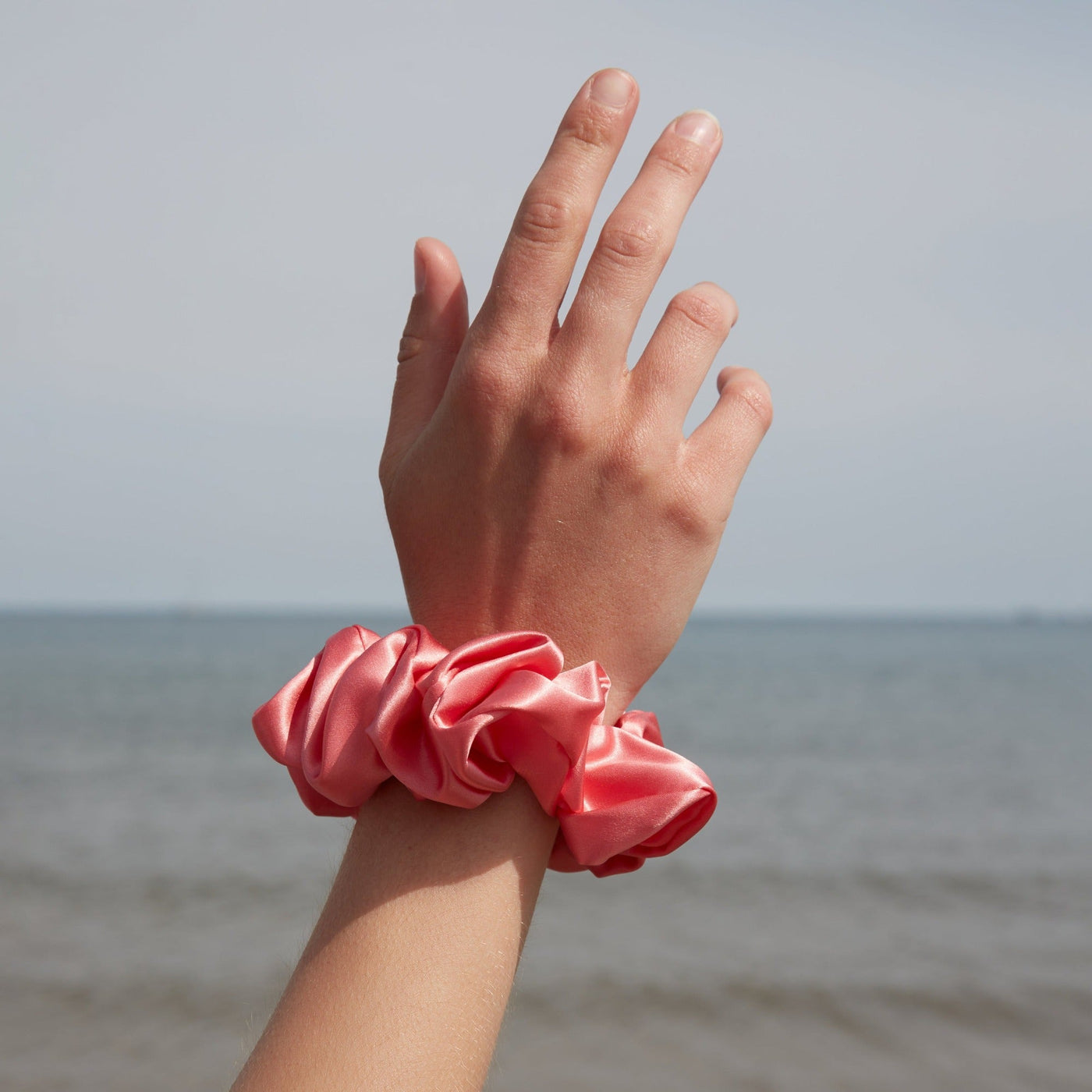 Hand wearing a Silk Works large watermelon pink mulberry silk scrunchie, photographed against a seaside backdrop.