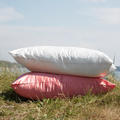 Stack of Silk Works mulberry silk pillowcases in ivory and watermelon pink, styled outdoors on grass under a bright summer sky.